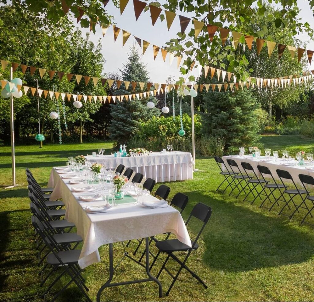 Outdoor event setup featuring elegant tables covered with white linens, decorated with floral centerpieces, and paired with sleek black folding chairs. The space is enhanced with decorative banners and hanging paper lanterns for a festive touch.