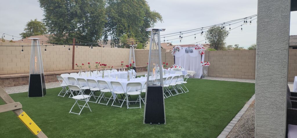 Outdoor event setup featuring elegant white tables and chairs, decorated with red roses, string lights, and pyramid patio heaters, creating a warm and romantic atmosphere for a backyard celebration